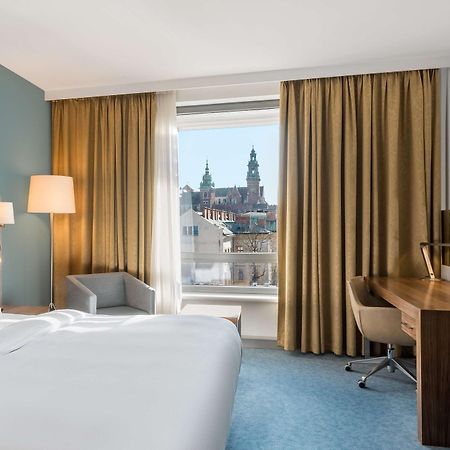Radisson Blu Hotel Krakow Exterior photo The photo depicts a hotel room featuring a large bed with white linens, a light-colored headboard, and a side lamp. There is a comfortable armchair in the corner, and a wooden desk with a chair positioned near the window. The window reveals a view of