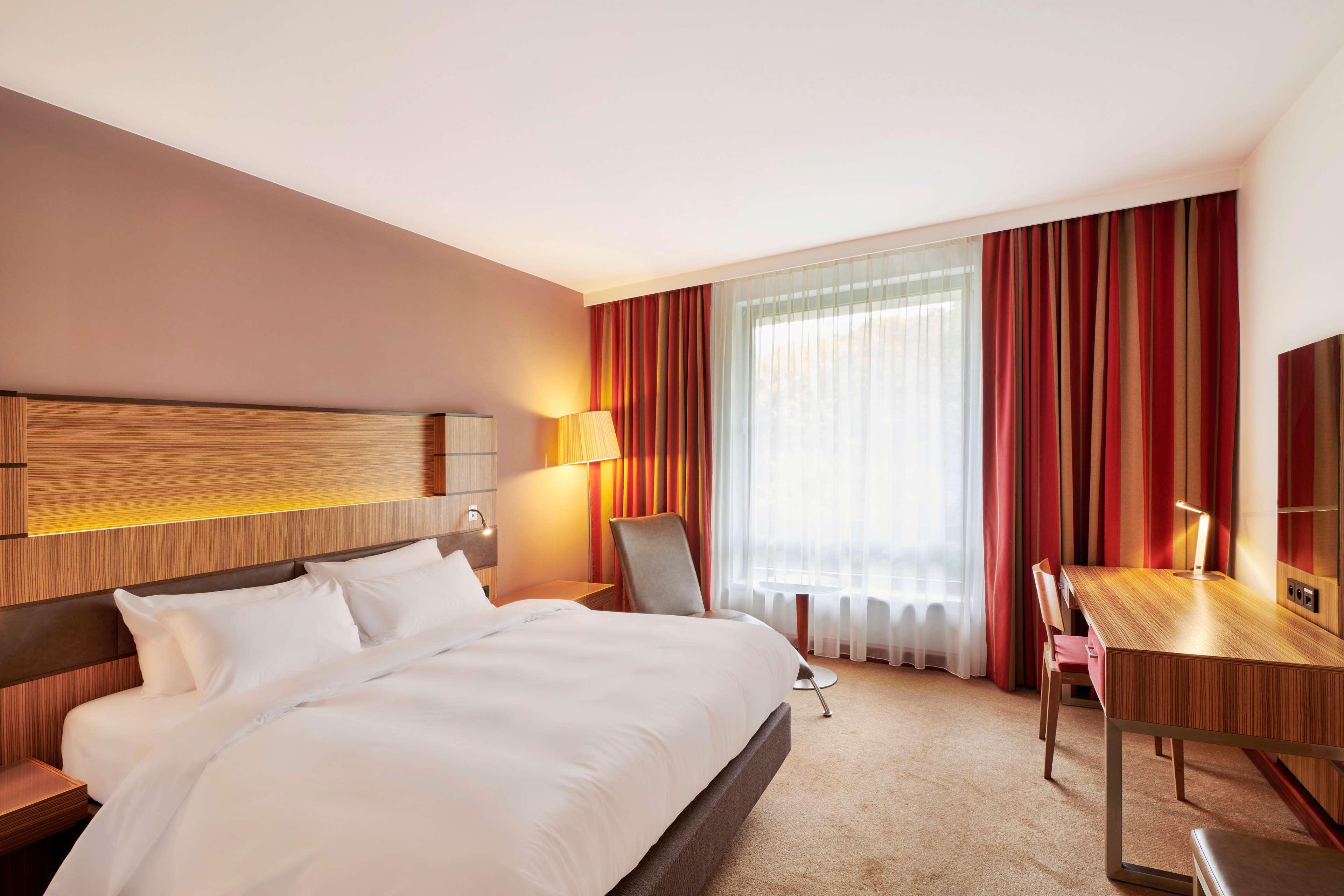 Radisson Blu Hotel Krakow Exterior photo The photo shows a modern hotel room. It features a large bed with white bedding against a wall that has wooden accents. There's a bedside lamp and a comfortable chair nearby. A window with sheer curtains allows natural light to enter, and the room ha