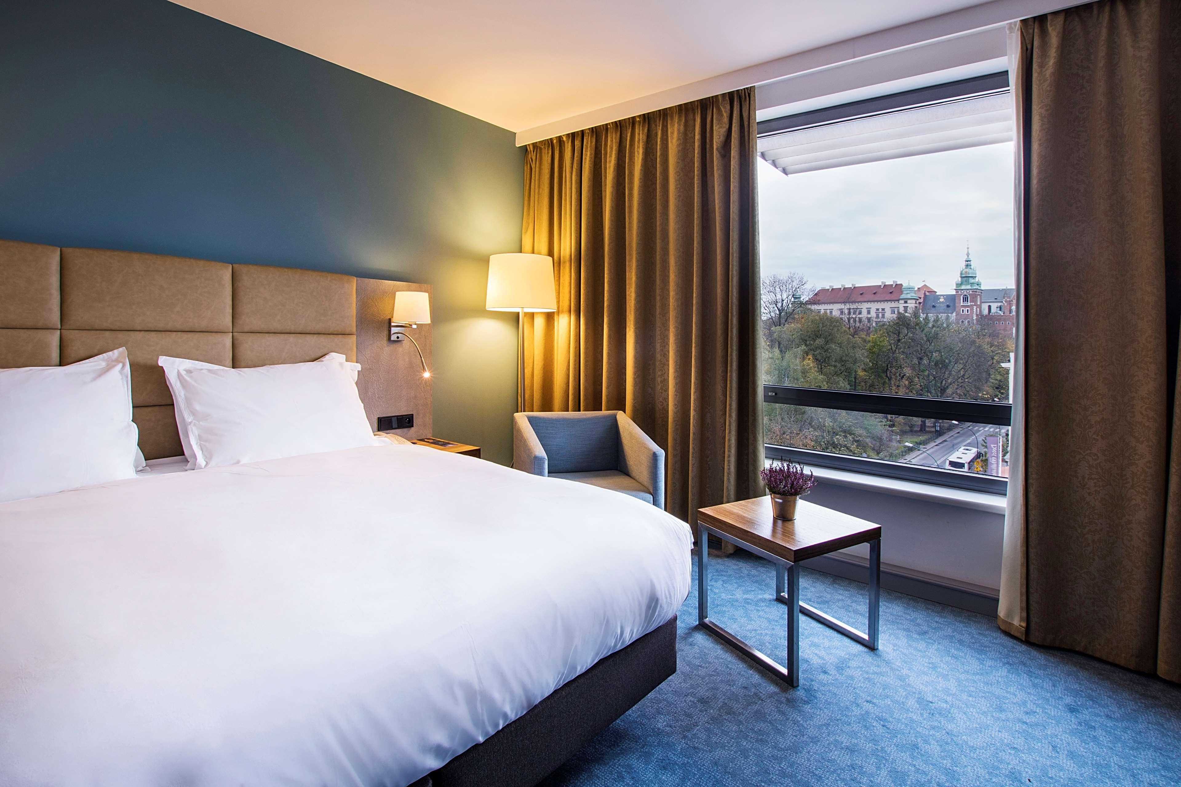 Radisson Blu Hotel Krakow Exterior photo The photo shows a modern hotel room featuring a queen-size bed with white bedding and two pillows. There is a bedside lamp and a small armchair in the corner next to a window. The window has grey curtains, partially open, revealing a view of an exter
