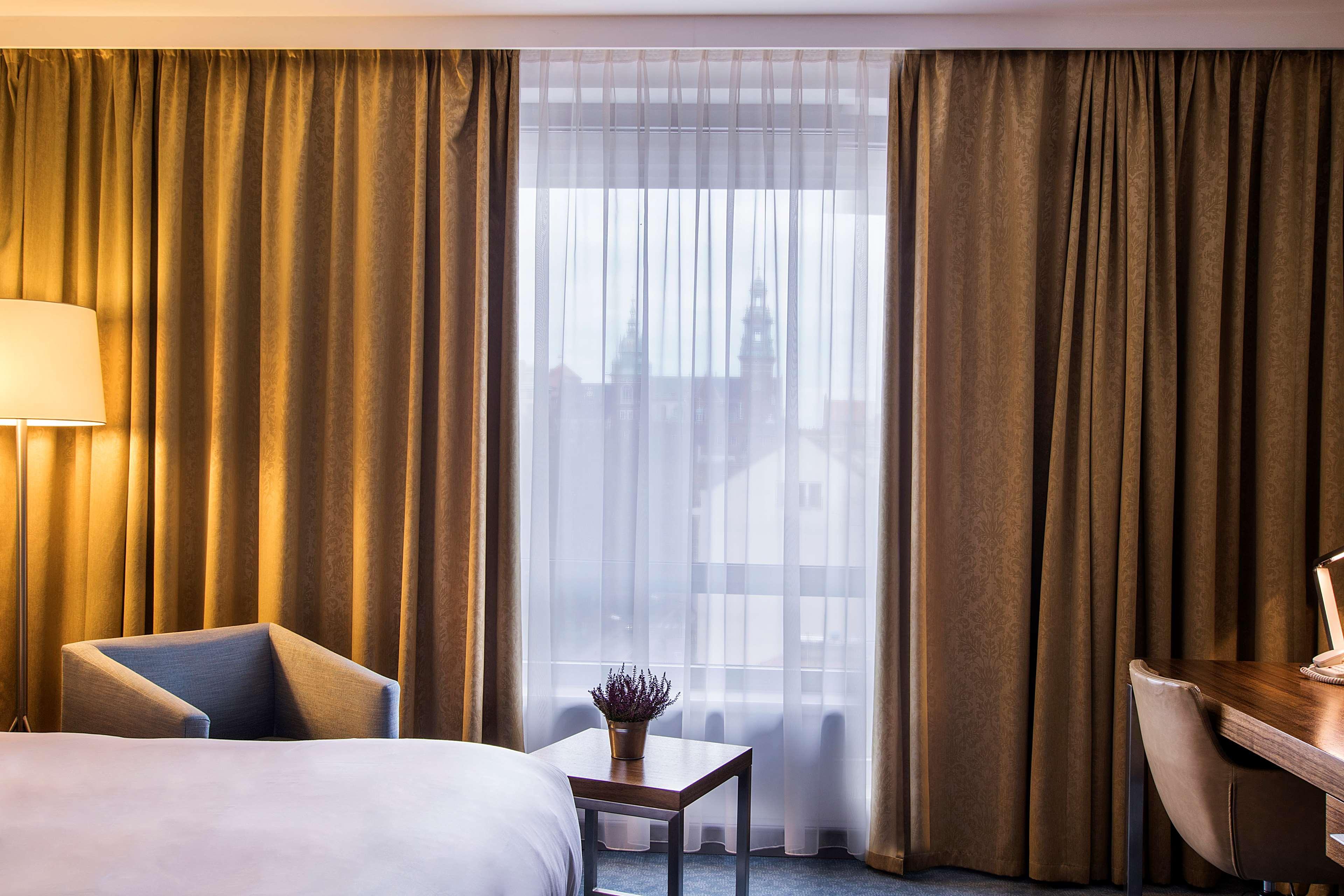 Radisson Blu Hotel Krakow Exterior photo The photo shows a modern hotel room with a soft color palette. On the left, there is a comfortable armchair beside a small table with a decorative plant. The room has large windows covered with sheer curtains and gold-colored drapes, allowing soft li