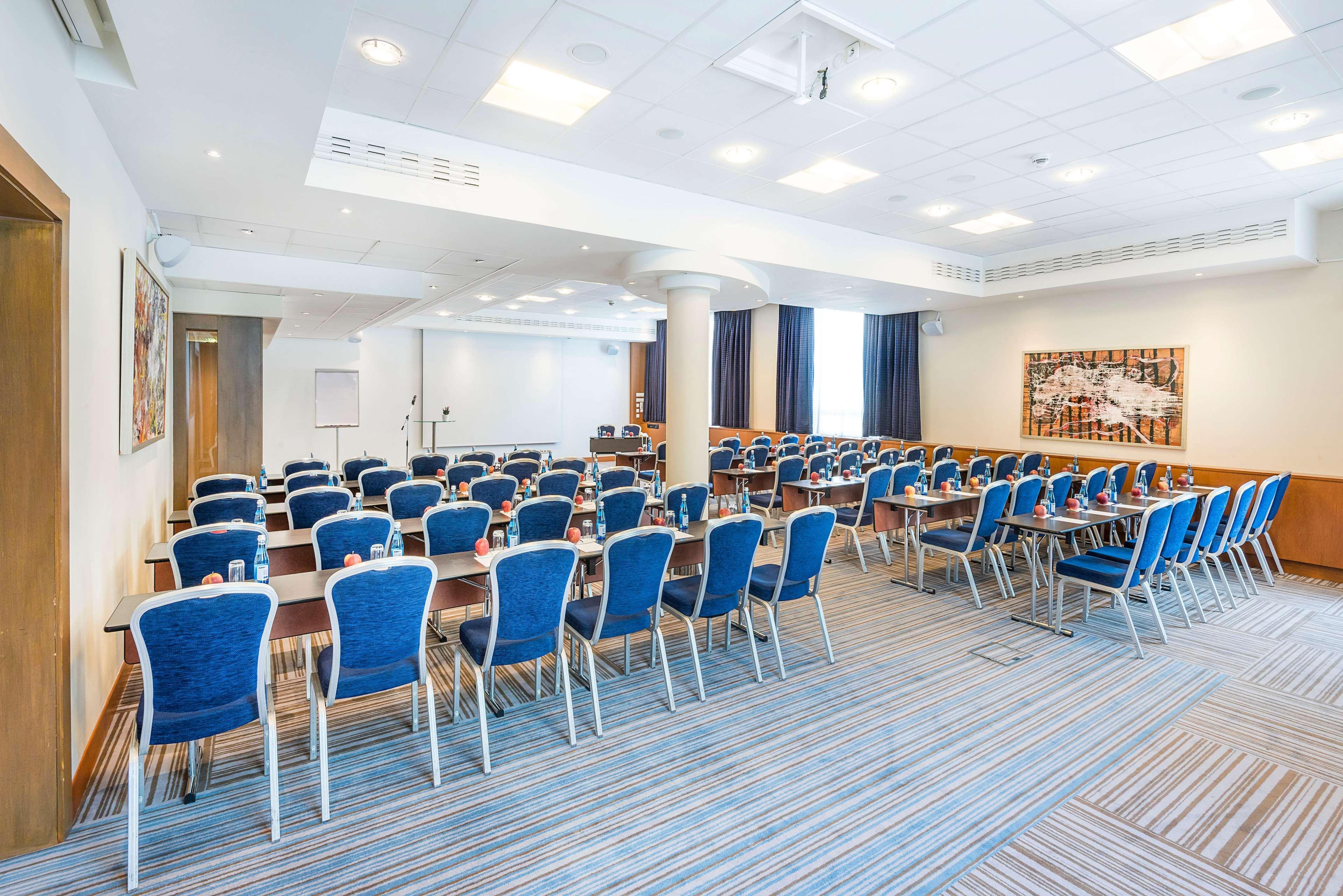Radisson Blu Hotel Krakow Exterior photo The photo shows a meeting or conference room set up for attendees. Rows of blue chairs are arranged around wooden tables, providing seating for participants. The room has a modern design with bright lighting, carpeting featuring a striped pattern, an
