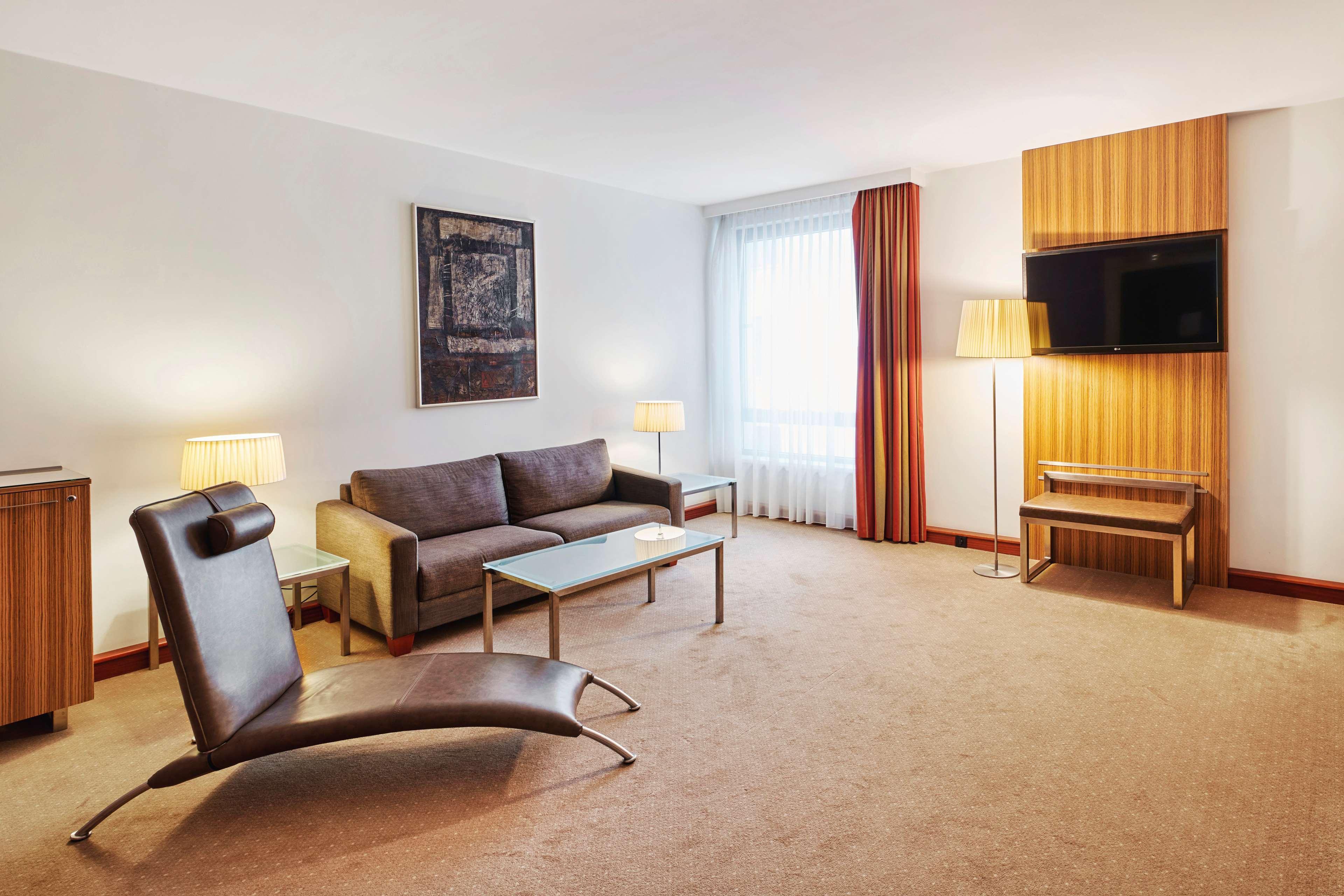 Radisson Blu Hotel Krakow Exterior photo The photo shows a modern hotel room or lounge area. It features a comfortable gray sofa accompanied by a glass coffee table. There is a distinctive brown, stylish lounge chair, adding a unique design element to the space. The room is softly lit with 