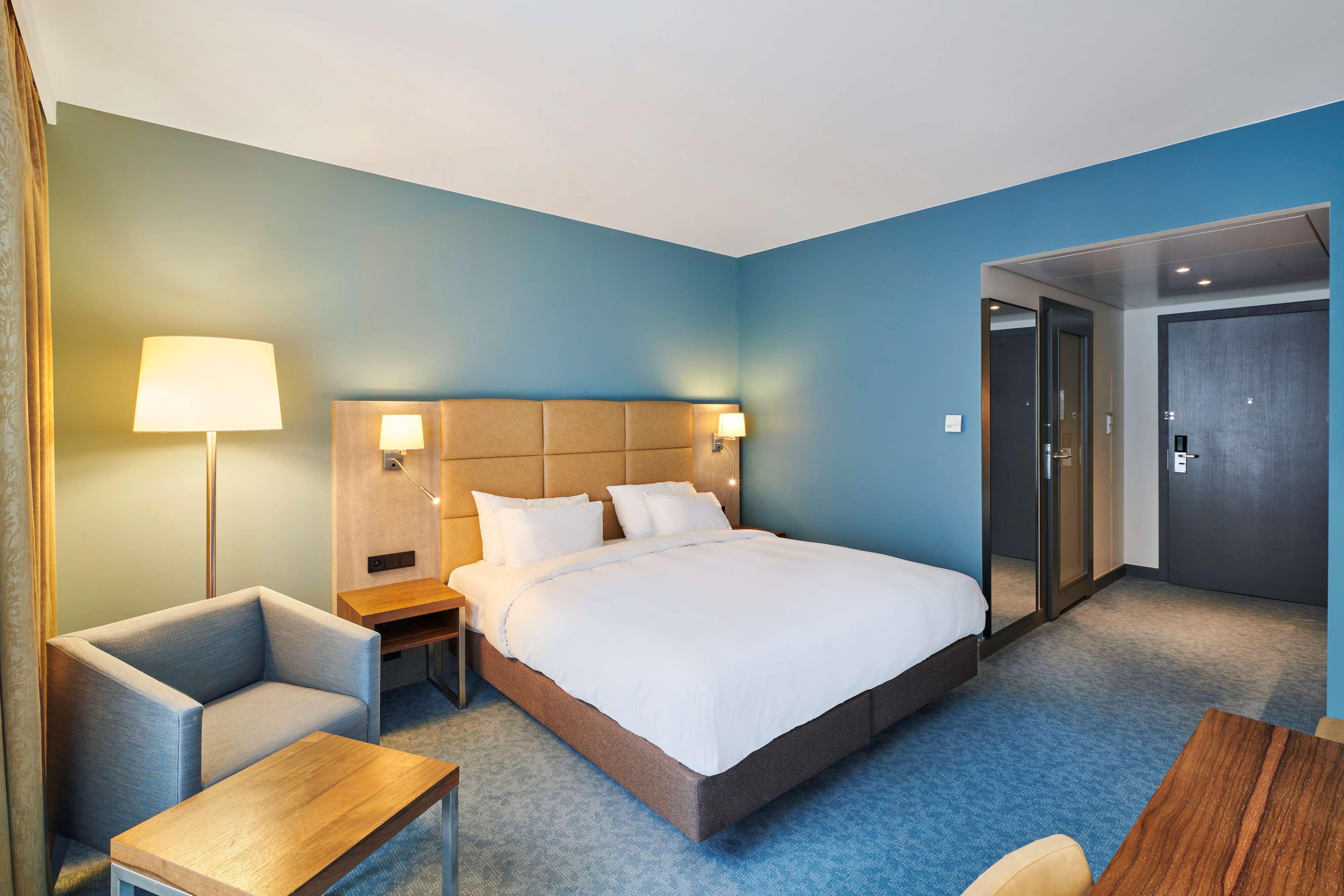 Radisson Blu Hotel Krakow Exterior photo The photo shows a modern hotel room featuring a queen-sized bed with white linens and decorative pillows. There are bedside lamps on either side of the bed, and a light blue accent wall adds to the room's contemporary decor. To the side, there is a s