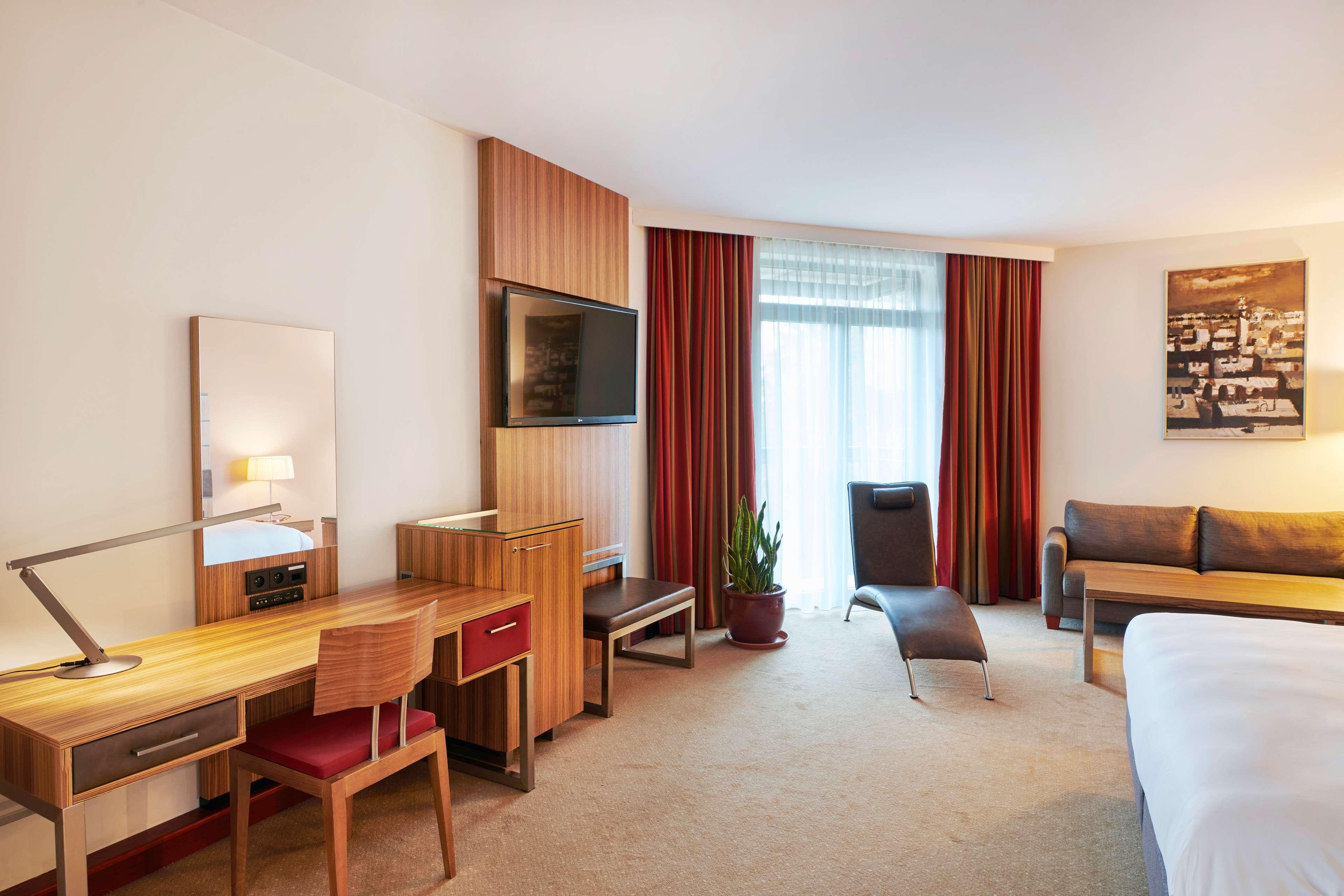 Radisson Blu Hotel Krakow Exterior photo The photo shows a hotel room that features a modern and comfortable layout. It includes a double bed, a seating area with a sofa, and a large window covered by sheer and dark curtains, allowing natural light to enter the space. There is a wooden desk