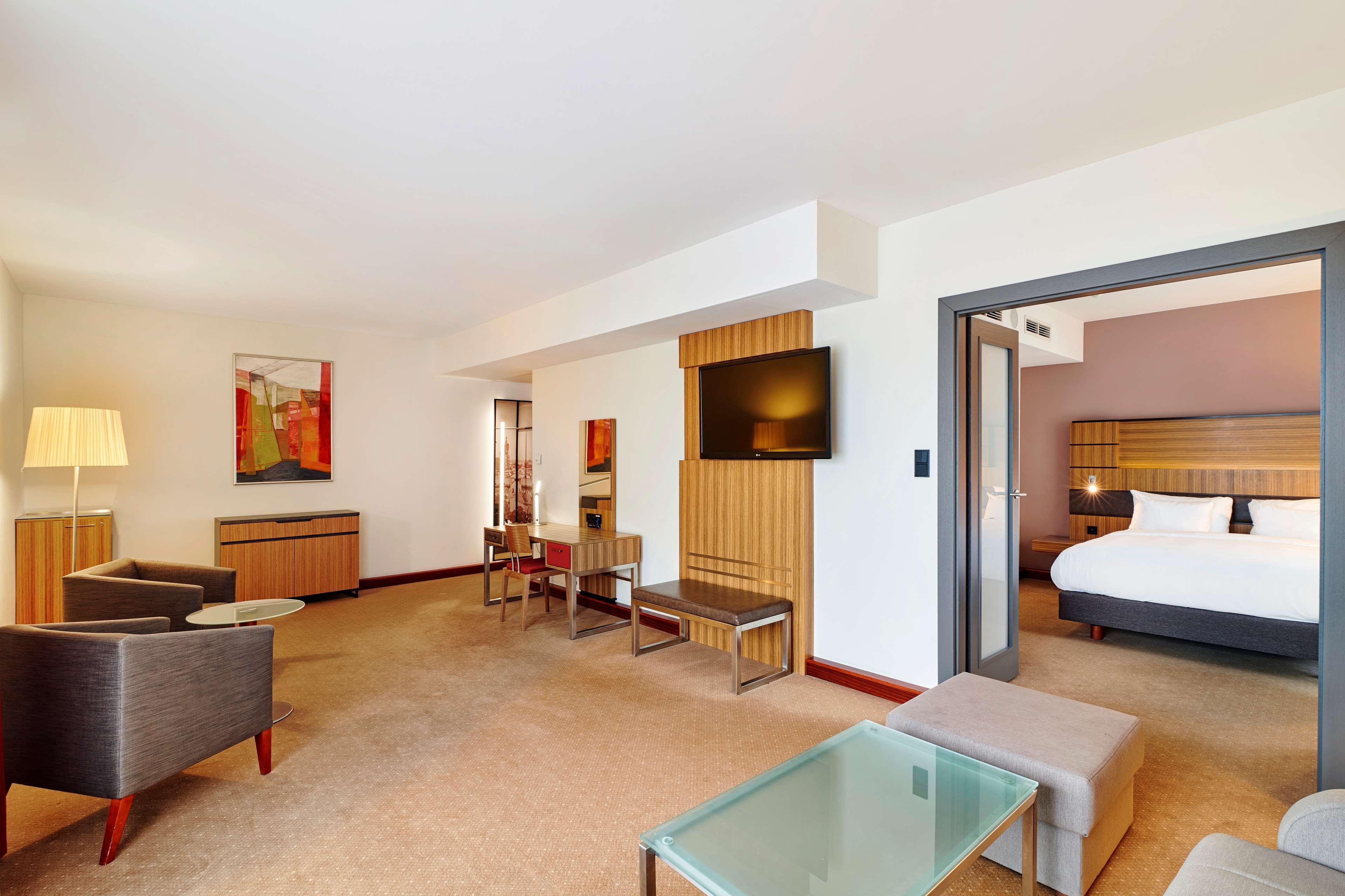 Radisson Blu Hotel Krakow Exterior photo The photo shows a hotel room or suite that features a spacious and modern design. On one side, there are two gray armchairs accompanied by a small table, creating a cozy seating area. A glass-top table and some additional seating can also be seen.

I