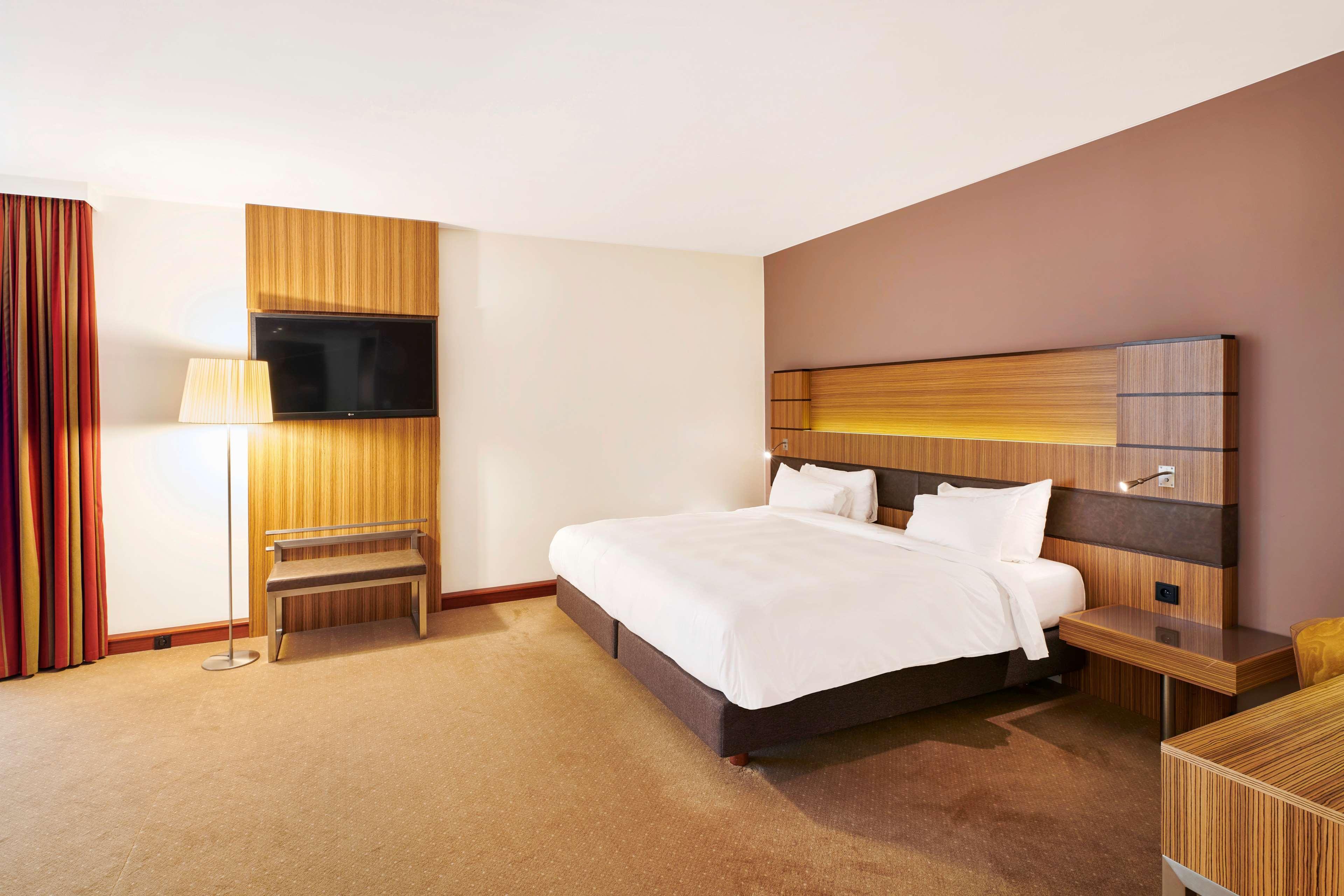 Radisson Blu Hotel Krakow Exterior photo The photo shows a modern hotel room featuring a large double bed with white bedding and pillows. There is a wooden headboard with integrated lighting above the bed. On the wall opposite the bed, a flat-screen television is mounted. To the side, there