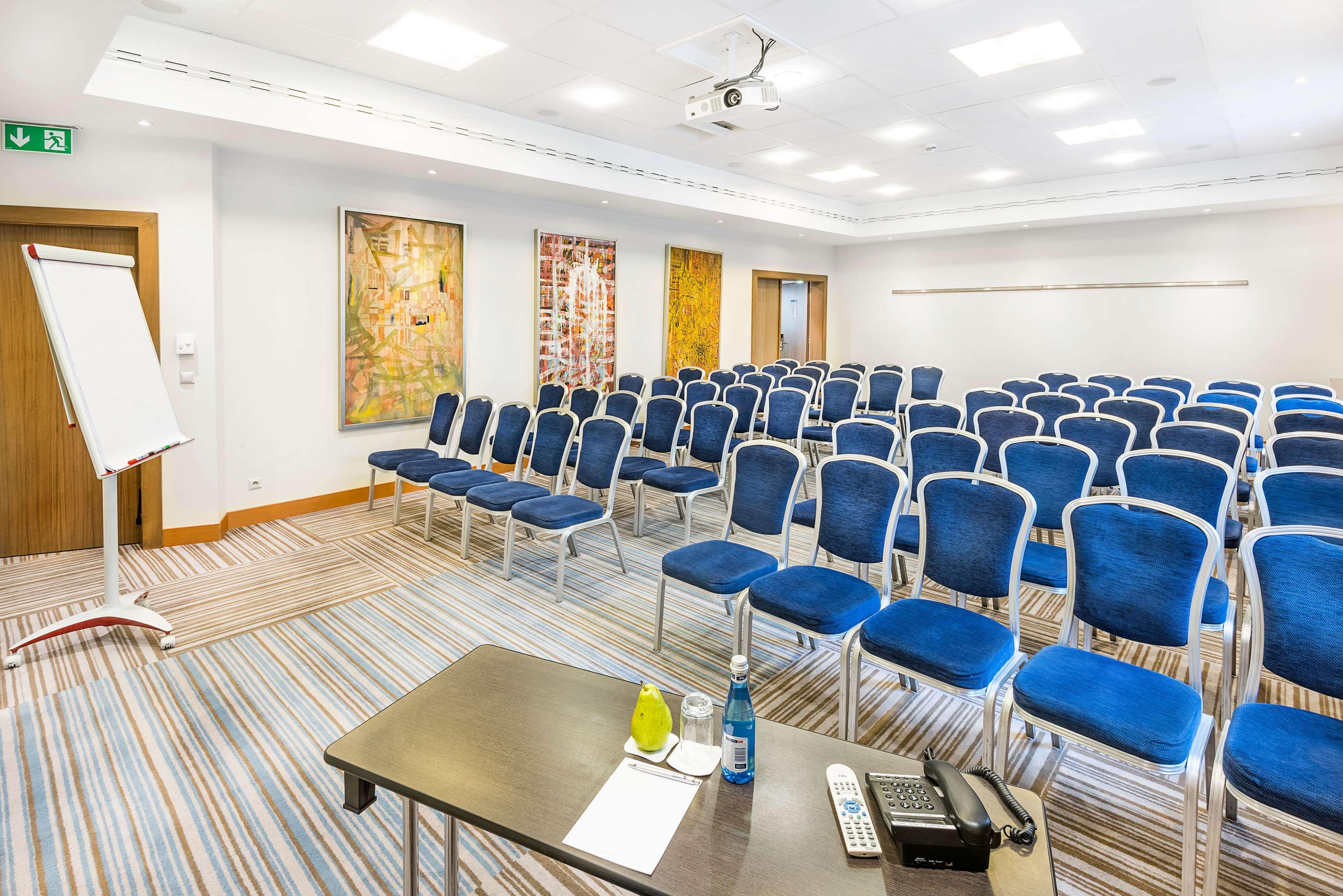 Radisson Blu Hotel Krakow Exterior photo The photo shows a conference room set up for an event or meeting. There are multiple rows of blue chairs arranged in a semi-theater style, facing a presentation area. In the foreground, there is a table with a few items, including a bottle of water a