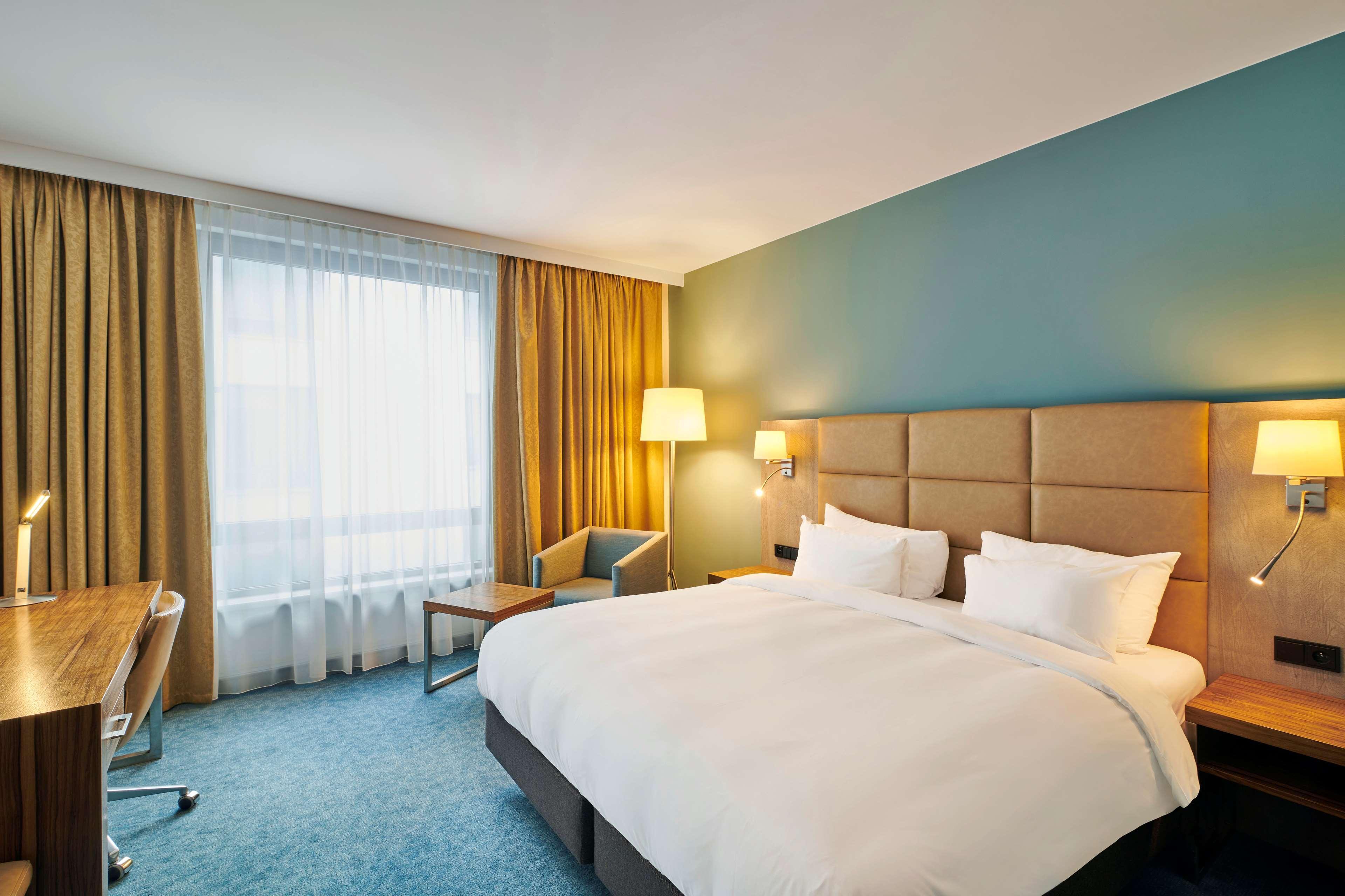 Radisson Blu Hotel Krakow Exterior photo The photo shows a modern hotel room. It features a large bed with white bedding and pillows. The walls are painted a calming blue, and there are large windows covered with sheer curtains, allowing natural light to enter. Next to the bed, there are be