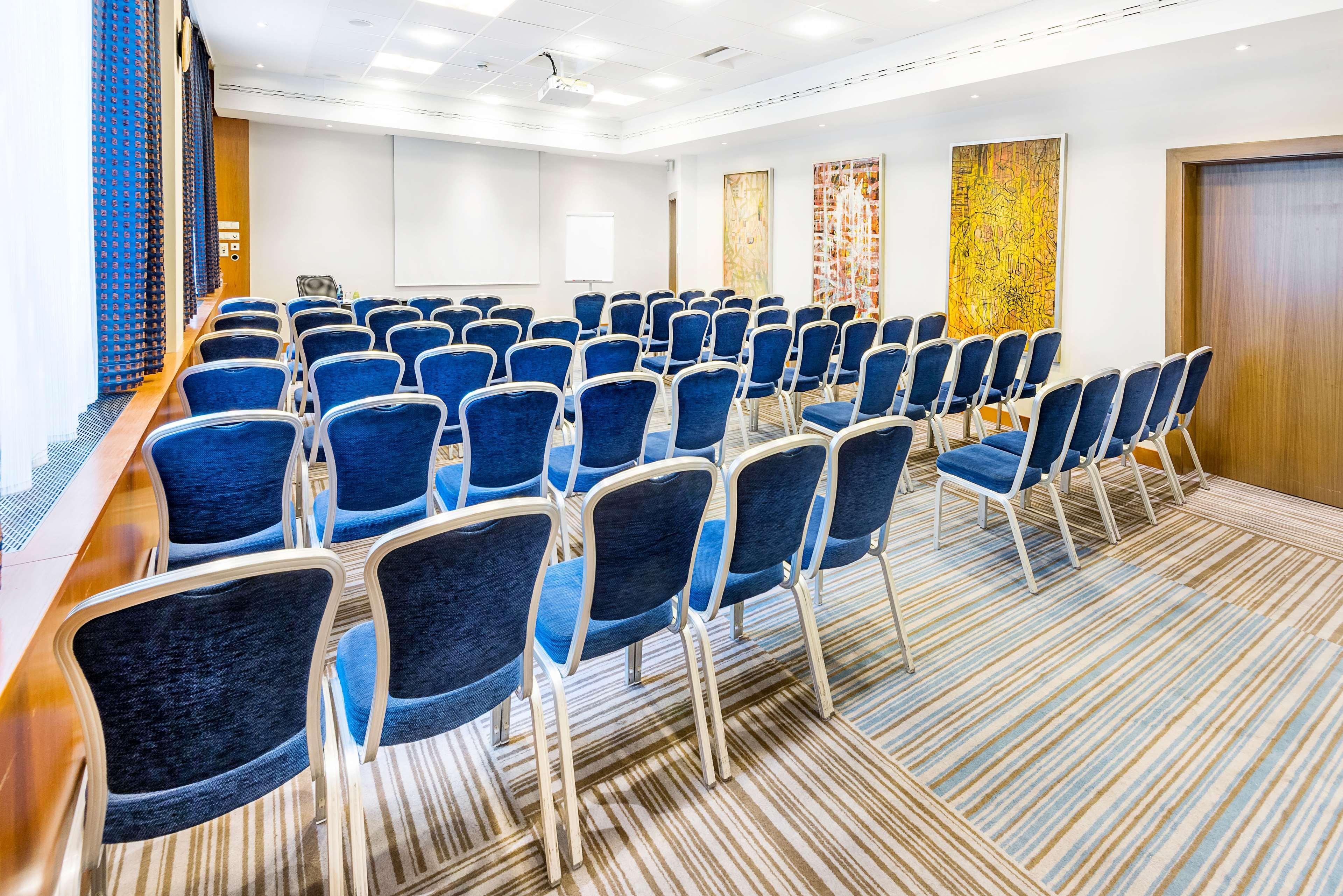 Radisson Blu Hotel Krakow Exterior photo The photo shows an indoor conference room set up for a presentation or meeting. There are several rows of blue chairs arranged facing the front of the room. The chairs have silver metal frames and plush cushions. On the walls, there are decorative pa
