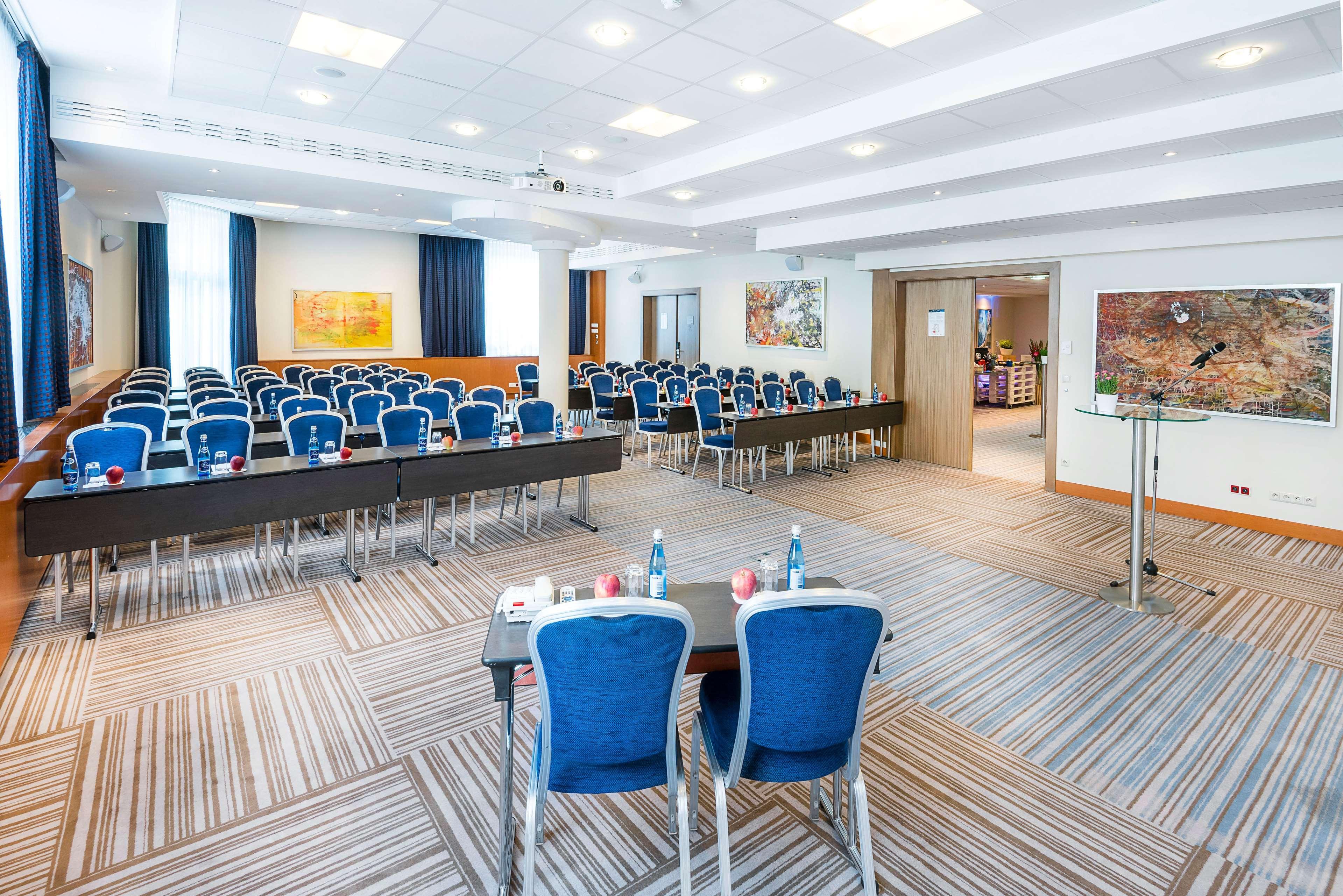 Radisson Blu Hotel Krakow Exterior photo The photo shows a conference room set up for a meeting or seminar. It features multiple rows of blue chairs arranged behind long tables. The room is well-lit with natural light coming from large windows, and there are decorative artworks on the walls