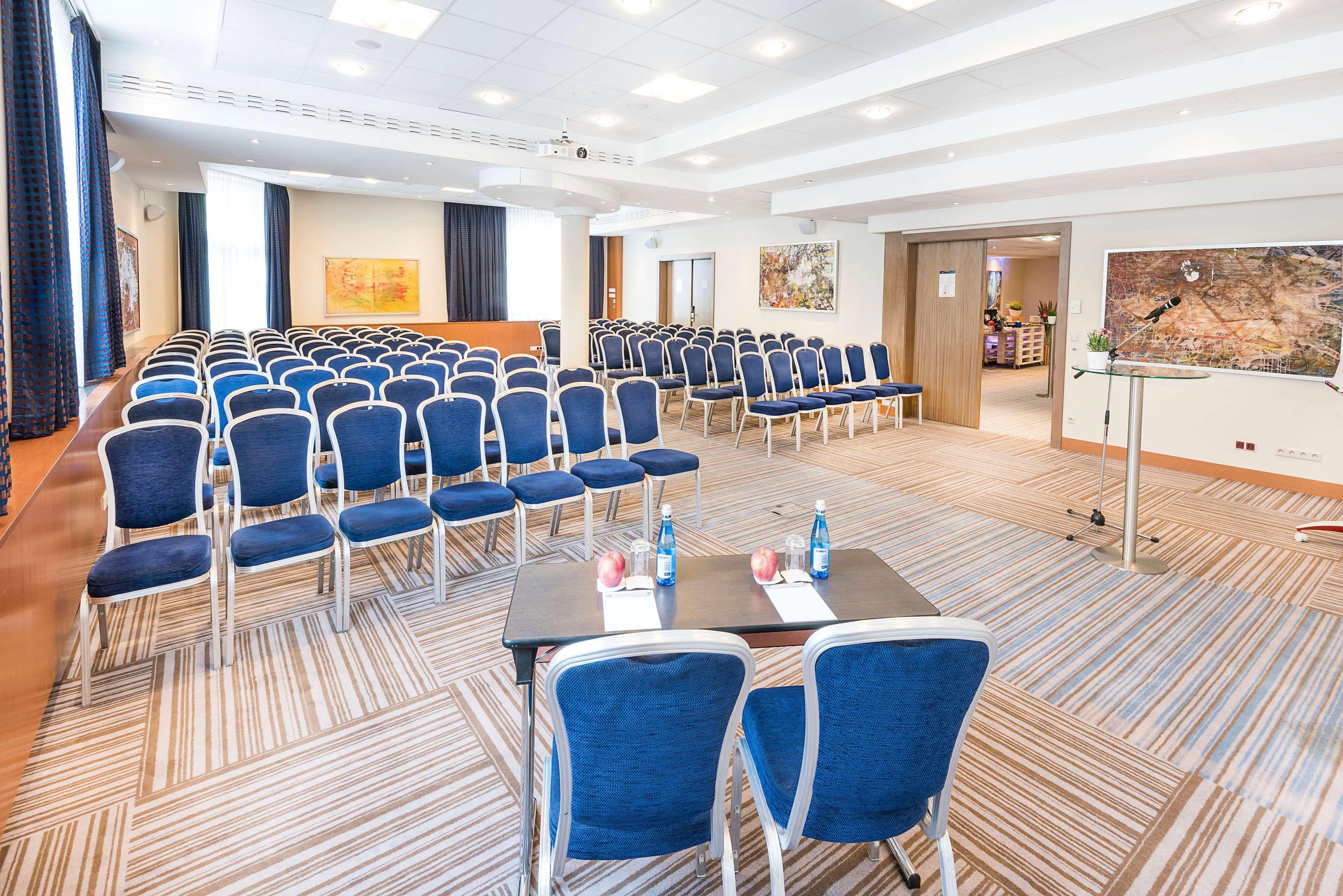 Radisson Blu Hotel Krakow Exterior photo The photo shows a conference room set up for an event. There are several rows of blue upholstered chairs arranged facing a small stage or presentation area. In the foreground, there are two tables with water bottles and some red decorations on them. 