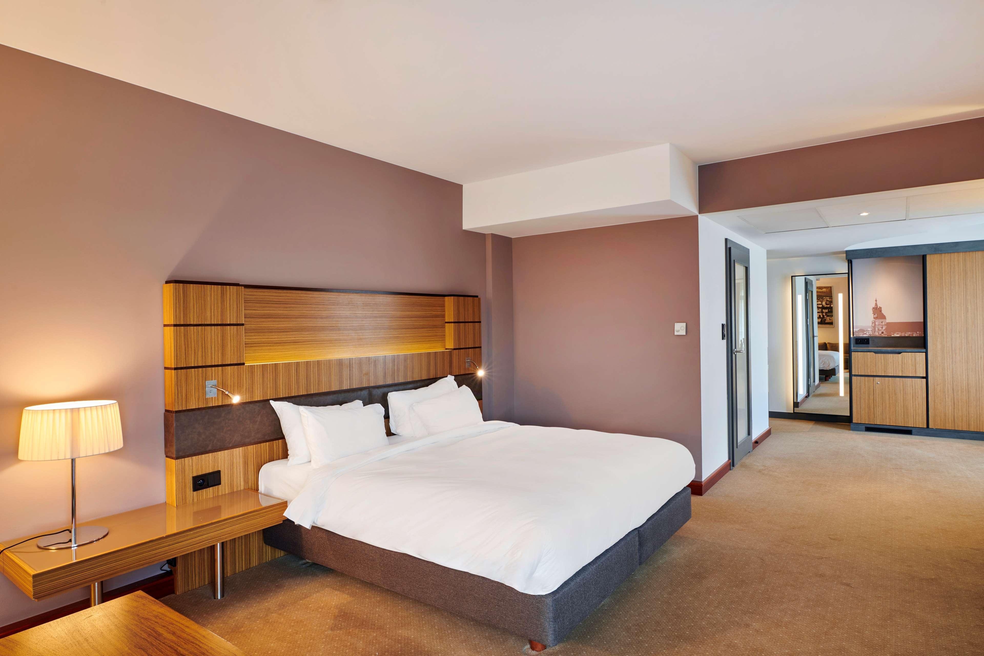 Radisson Blu Hotel Krakow Exterior photo The photo shows a modern hotel room featuring a double bed with white bedding and a gray upholstered base. There is a wooden headboard with built-in lighting. To one side of the bed, there's a small wooden table with a lamp. The room has a neutral co