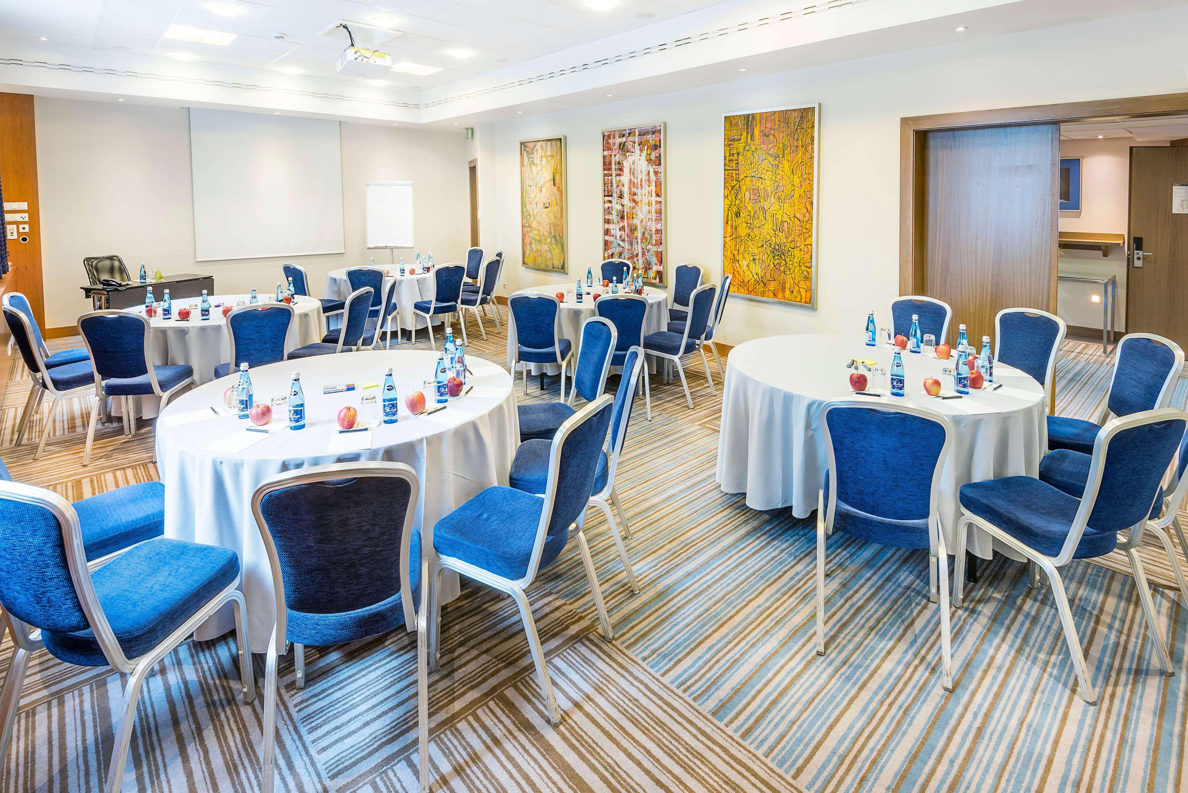 Radisson Blu Hotel Krakow Exterior photo The photo shows a conference or meeting room set up for an event. There are several round tables covered with white tablecloths, each accompanied by blue chairs. Bottled water and small notepads appear to be placed on the tables. The room features mo