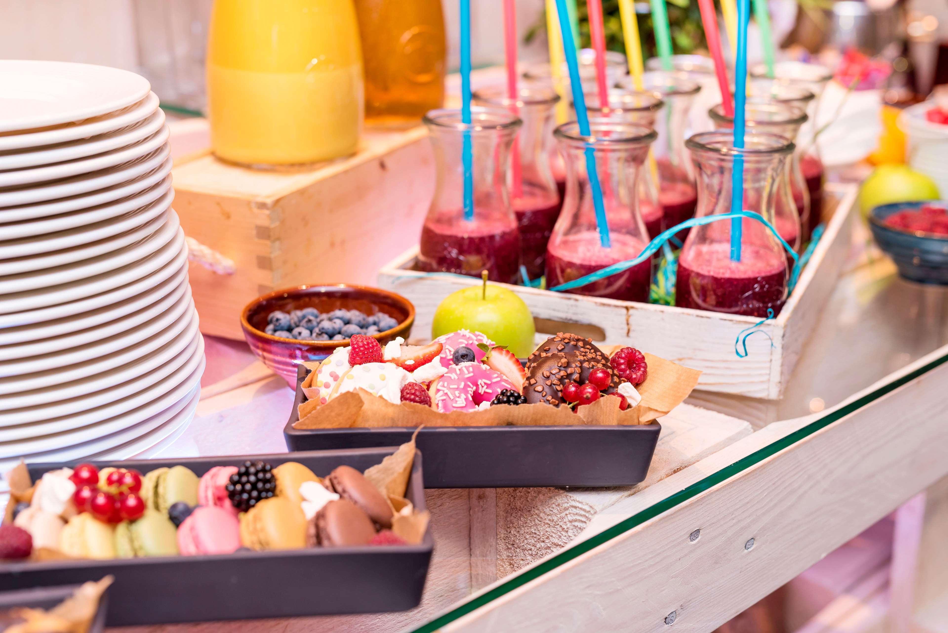Radisson Blu Hotel Krakow Exterior photo The photo features a vibrant display of desserts and drinks on a table. There are several stacked white plates on the left side. In the center, there are small glass bottles filled with a colorful smoothie or juice, each featuring a straw. A white wo