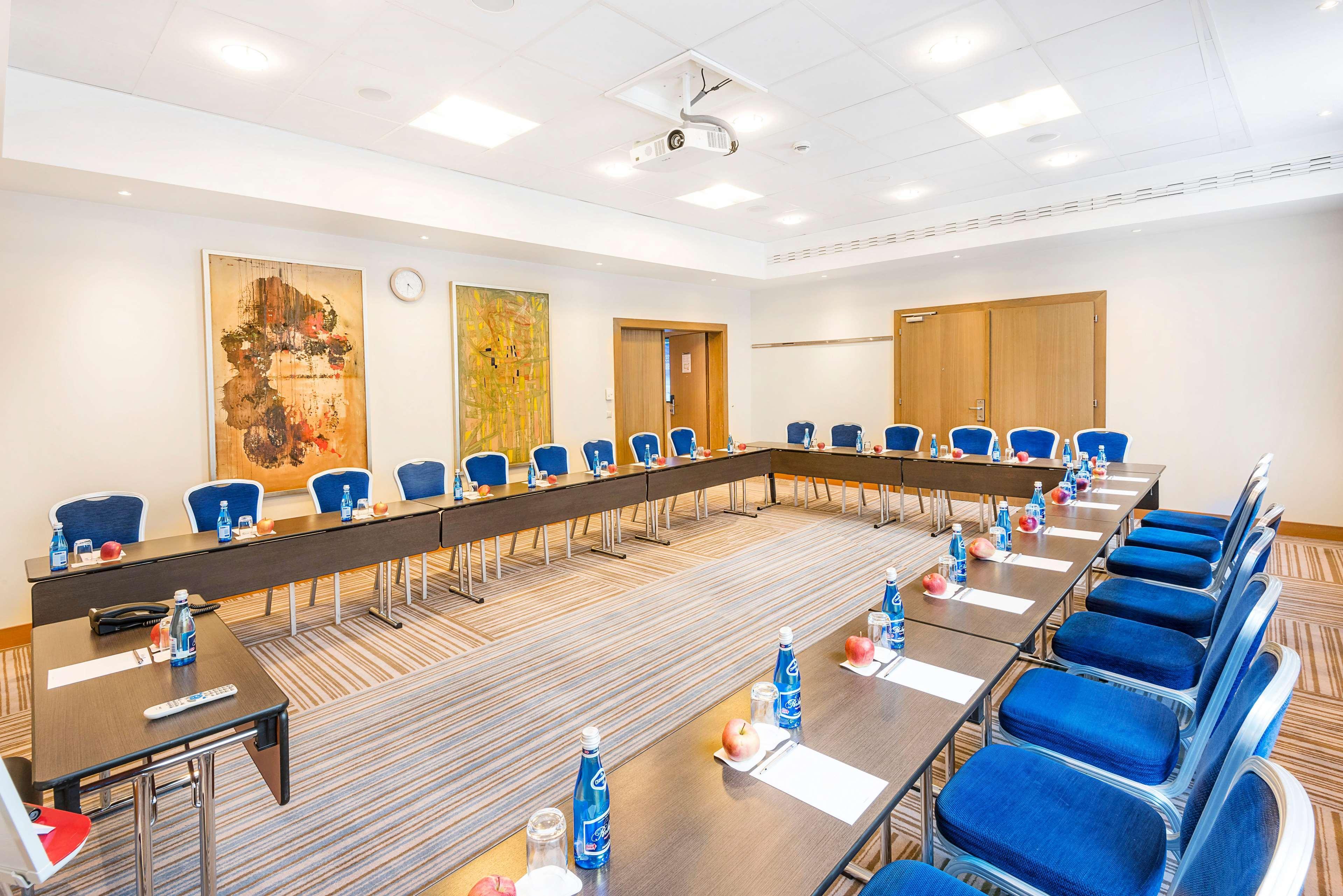 Radisson Blu Hotel Krakow Exterior photo The photo shows a meeting room set up for a conference or workshop. There are two long tables arranged in a U-shape, with blue chairs positioned around them. Each table is equipped with bottles of water and apples, suggesting refreshments for attende