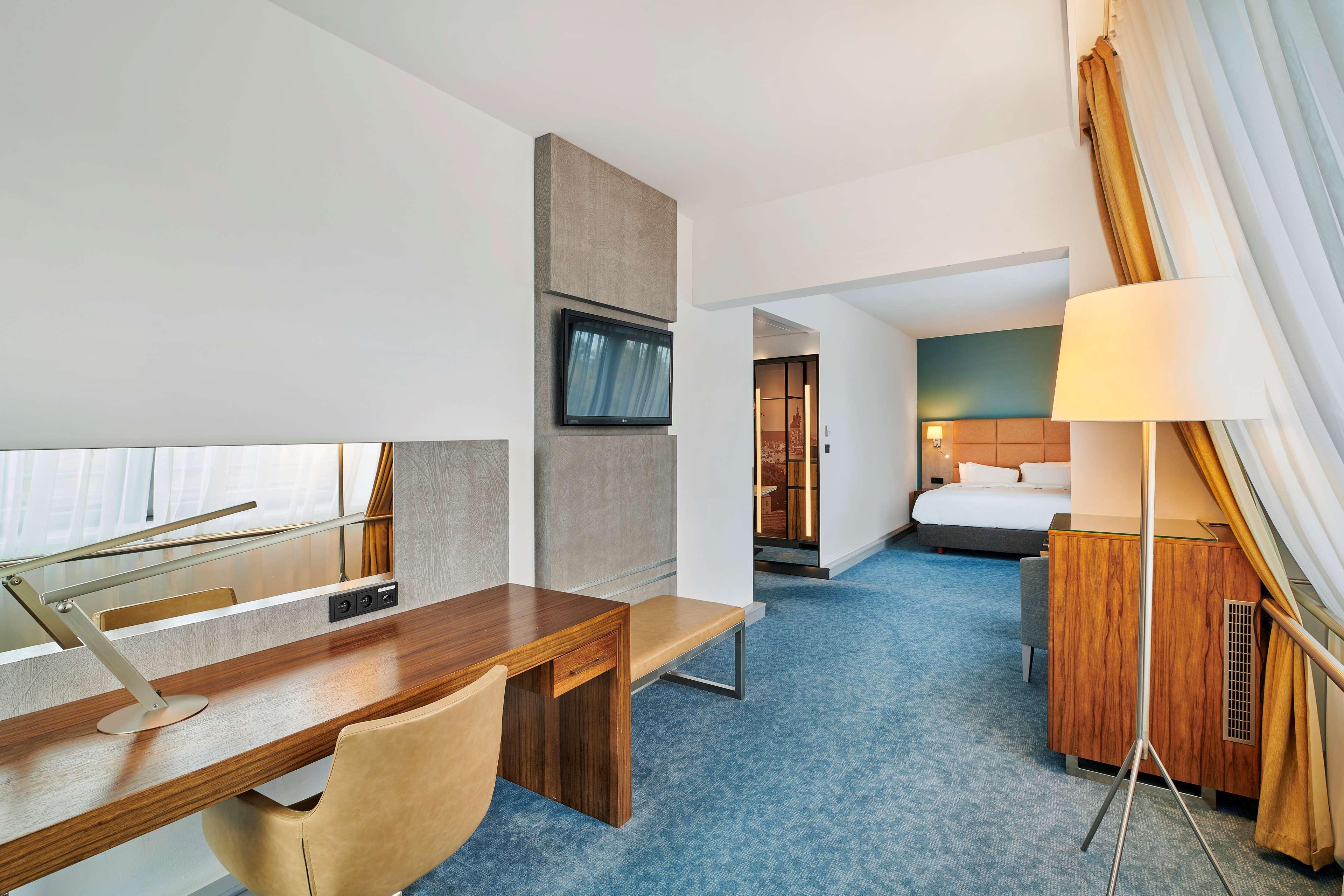 Radisson Blu Hotel Krakow Exterior photo The photo showcases a modern hotel room. On one side, there is a work desk made of wood with a stylish chair. The desk is accompanied by a large mirror and a wall-mounted TV above it. The flooring is carpeted in a soft blue color. In the background, 