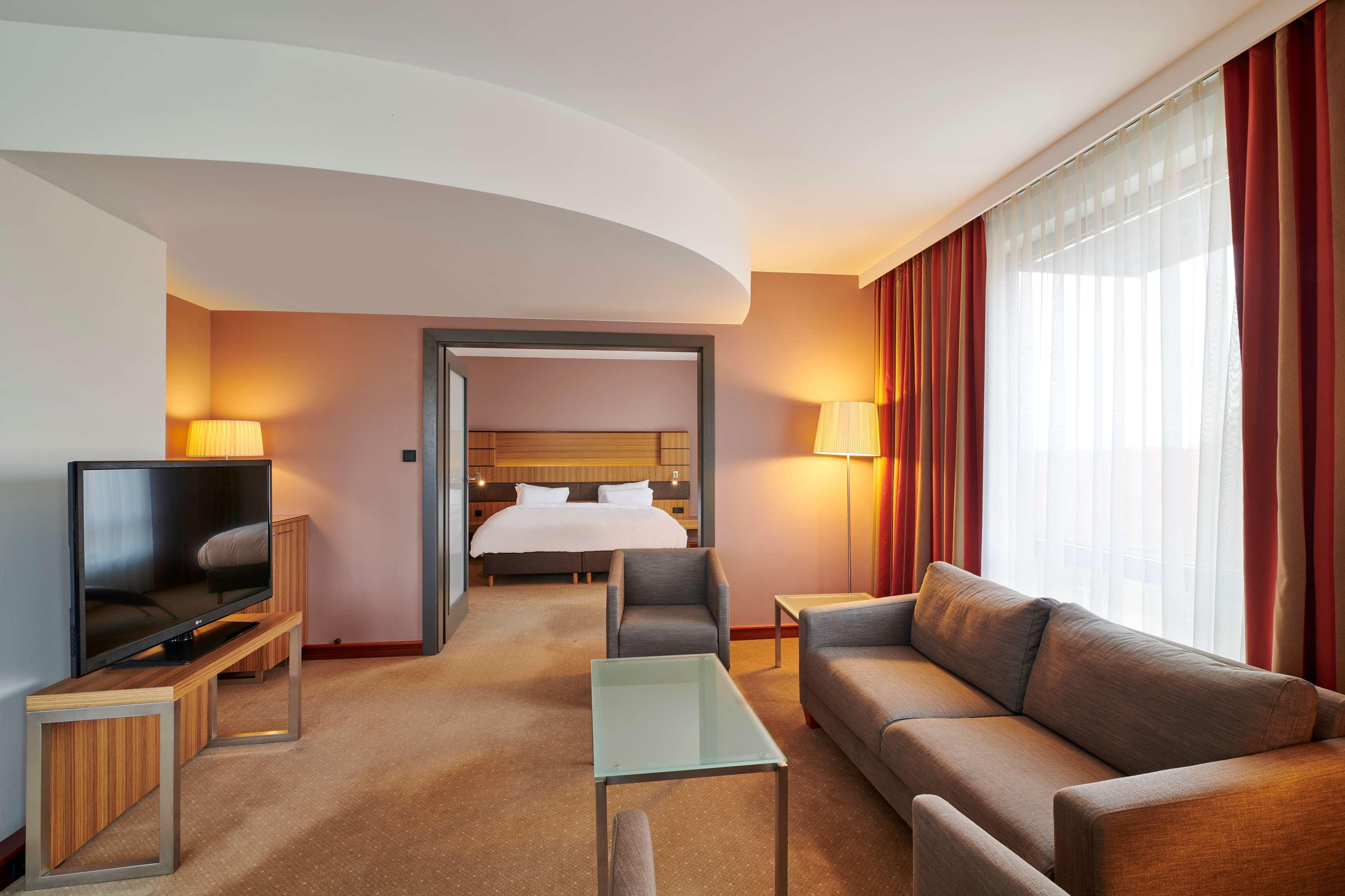 Radisson Blu Hotel Krakow Exterior photo The photo shows a modern hotel room suite. It features a cozy living area with a gray sofa, a glass coffee table, and a wooden TV stand with a flat-screen television. There are two lamps providing soft lighting. In the background, a door leads to the