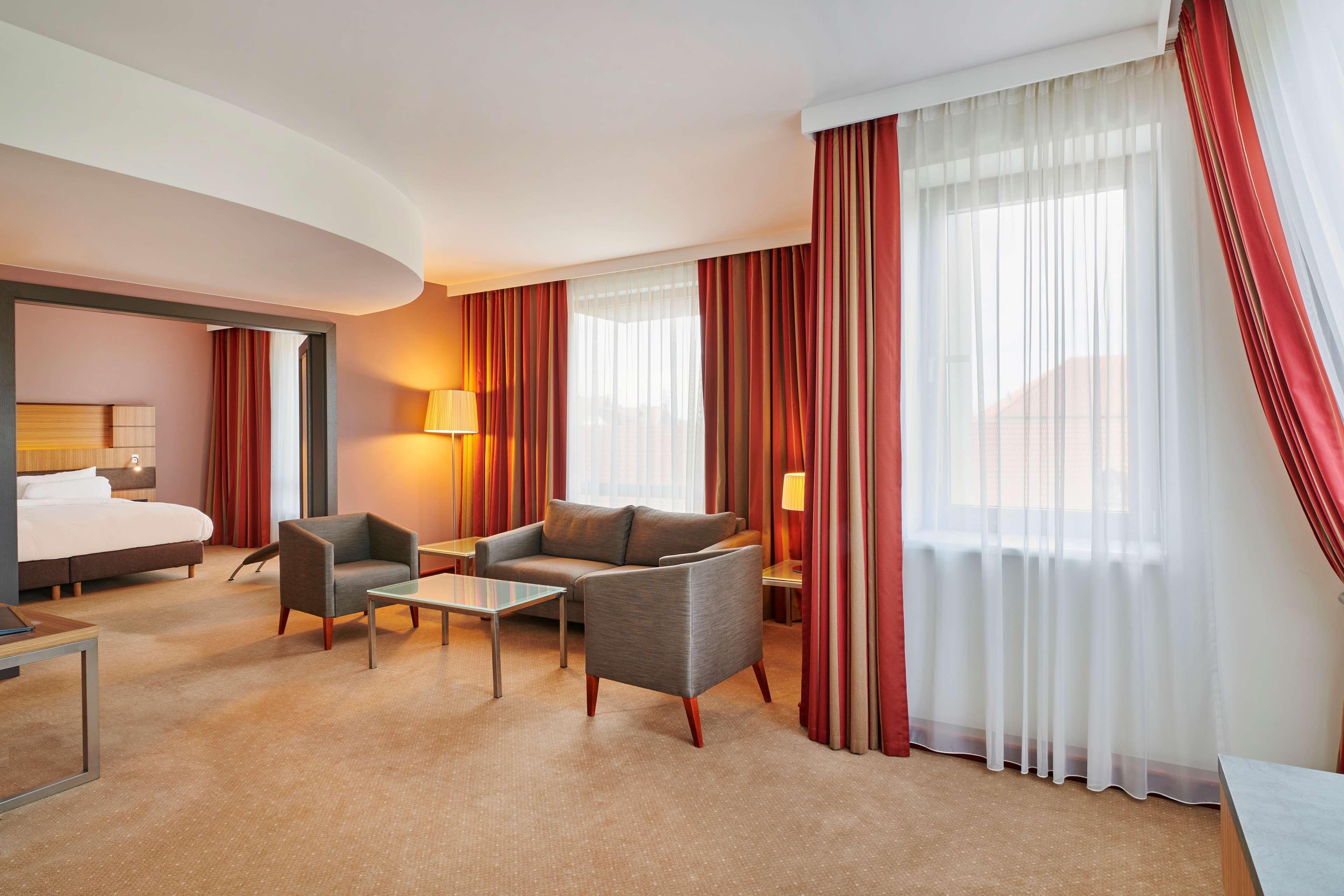 Radisson Blu Hotel Krakow Exterior photo The photo shows a well-furnished hotel room or suite. It features a cozy seating area with two gray armchairs and a small glass coffee table. A floor lamp provides additional lighting. The room is adorned with large windows dressed in sheer white cur