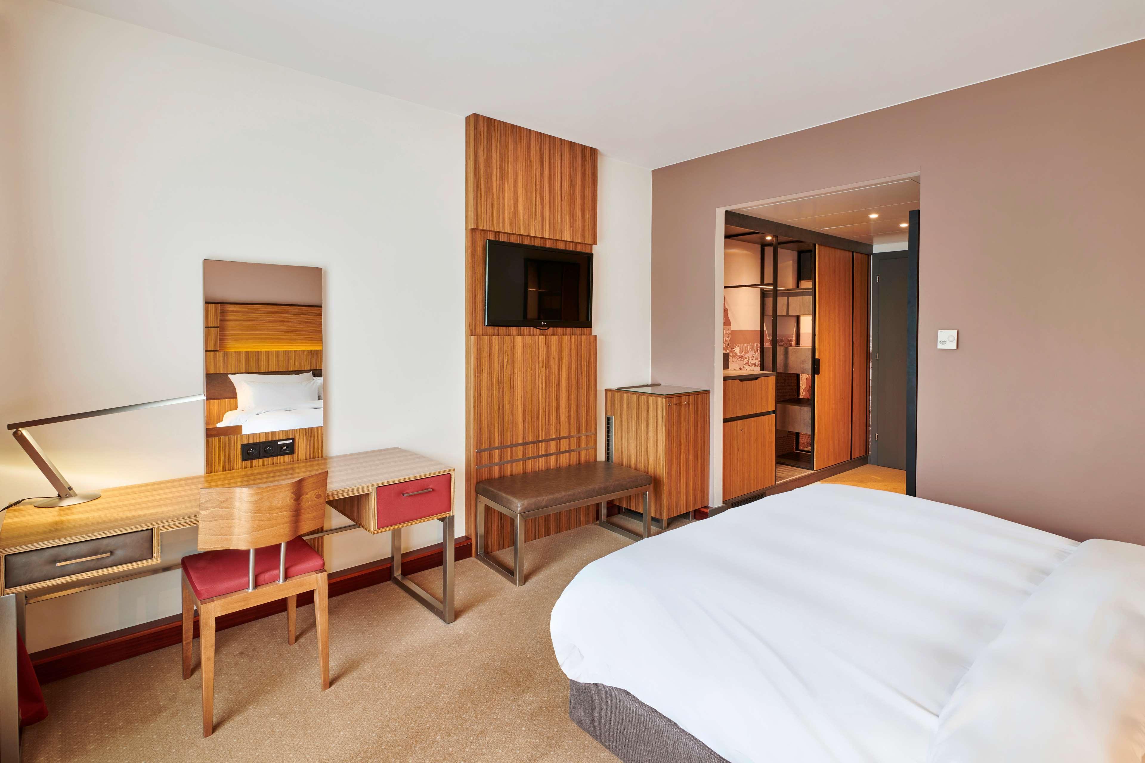 Radisson Blu Hotel Krakow Exterior photo The photo shows a modern hotel room with a minimalist design. It features a large bed with white bedding, positioned against a wall. To one side, there's a small seating area with a simple bench. Opposite the bed is a desk with a mirror and a chair, 