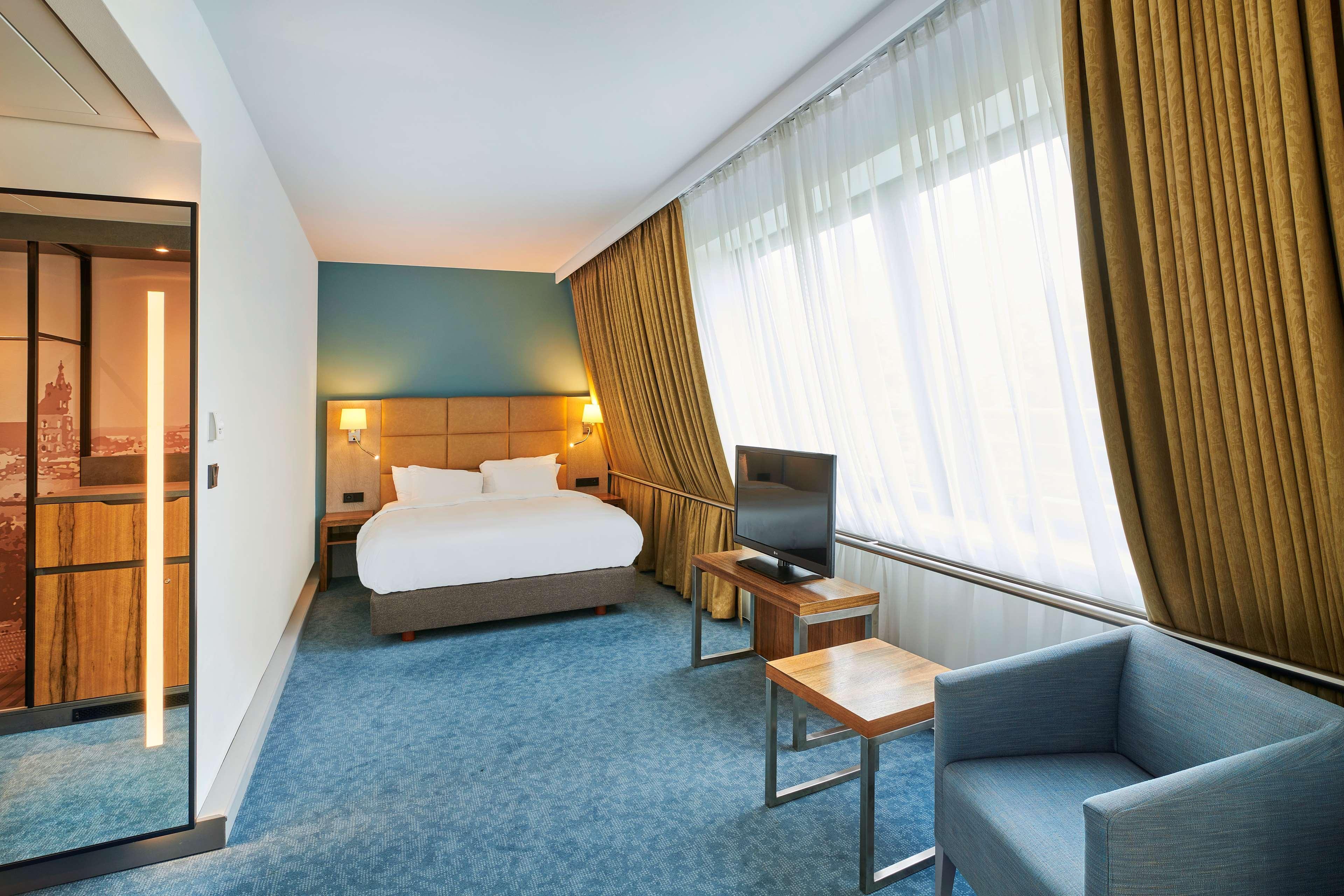 Radisson Blu Hotel Krakow Exterior photo The photo shows a modern hotel room. It features a queen-sized bed with white bedding, positioned against a wall with warm lighting. The room has a large window with sheer curtains that allow natural light to enter. There's a small wooden table and a
