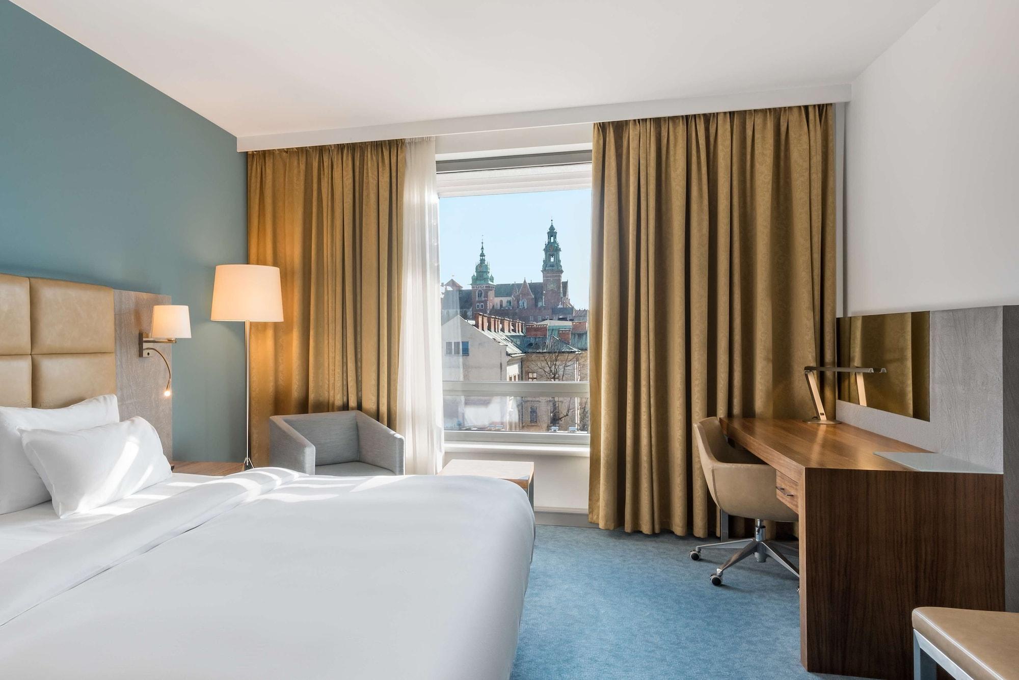 Radisson Blu Hotel Krakow Exterior photo The photo depicts a hotel room featuring a large bed with white linens, a light-colored headboard, and a side lamp. There is a comfortable armchair in the corner, and a wooden desk with a chair positioned near the window. The window reveals a view of