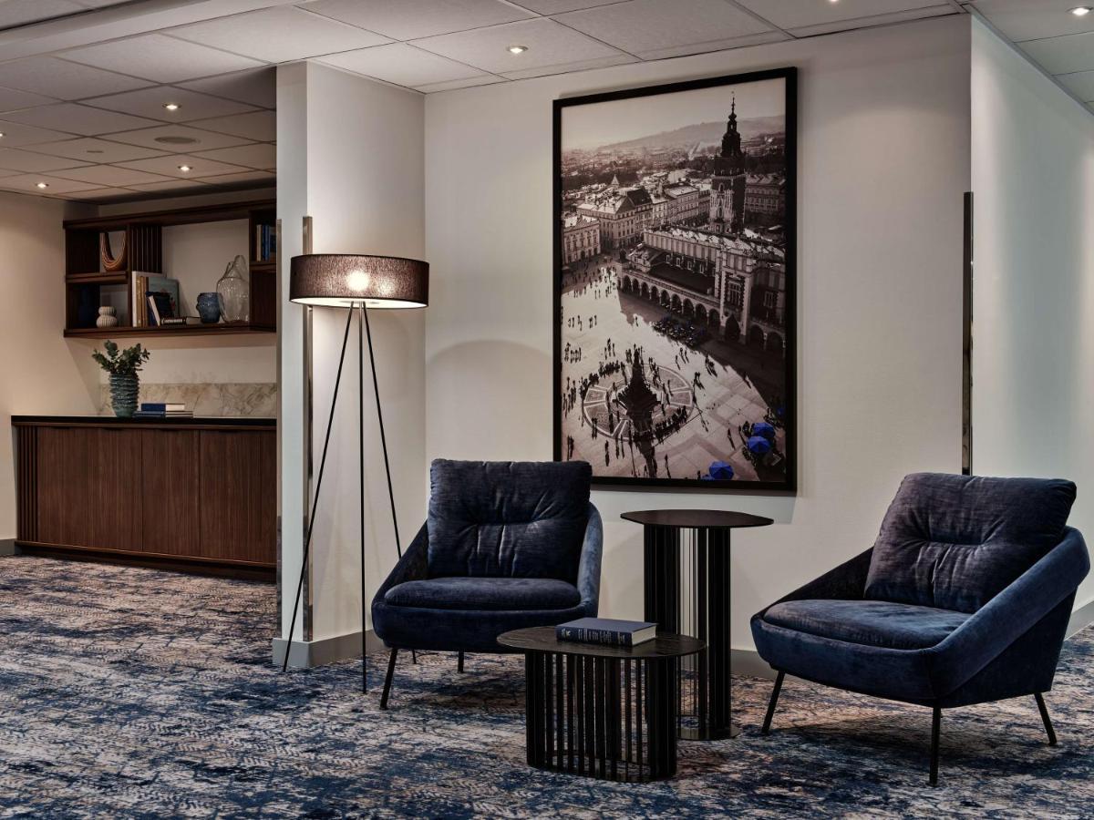 Radisson Blu Hotel Krakow Exterior photo The photo depicts a modern interior space that appears to be part of a hotel or a lounge area. In the foreground, there are two stylish blue armchairs accompanied by a small round black table. To the left, there is a reception desk with a decorative 
