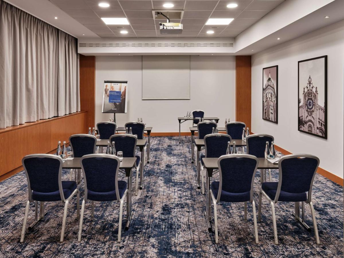 Radisson Blu Hotel Krakow Exterior photo The photo shows a conference room set up for a meeting or presentation. The room features a light-colored carpet with a patterned design. There are several rows of chairs arranged facing a small table, which has water bottles on it. A flip chart stan