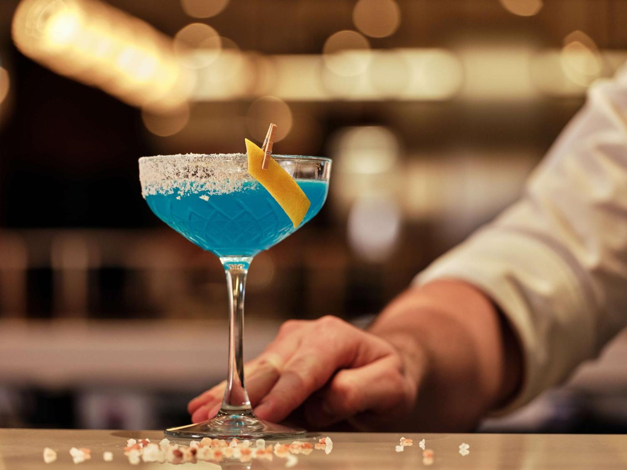 Radisson Blu Hotel Krakow Exterior photo The photo shows a vibrant blue cocktail served in a classic cocktail glass. The drink is garnished with a slice of lemon and has a rim coated with salt. In the background, there are blurred lights, creating a lively atmosphere, and a hand is gently p