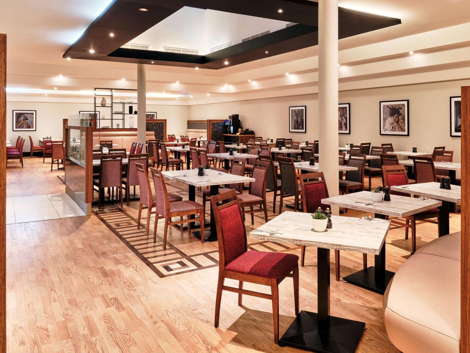 Radisson Blu Hotel Krakow Exterior photo The photo shows a spacious and modern restaurant interior. It features multiple tables surrounded by wooden chairs, some of which have red upholstery. The floor is made of light-colored wood, and there's a geometric pattern visible in the design. The