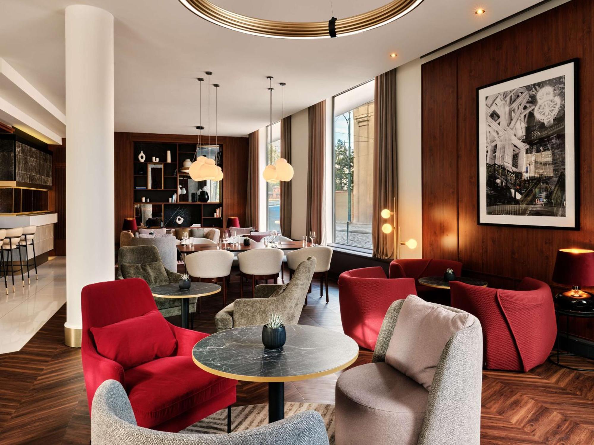 Radisson Blu Hotel Krakow Exterior photo The image depicts a stylish, modern interior of a lounge or café. The space features a mix of seating options, including red and gray armchairs arranged around small circular tables. The décor is warm and inviting, with wooden paneling on the walls a
