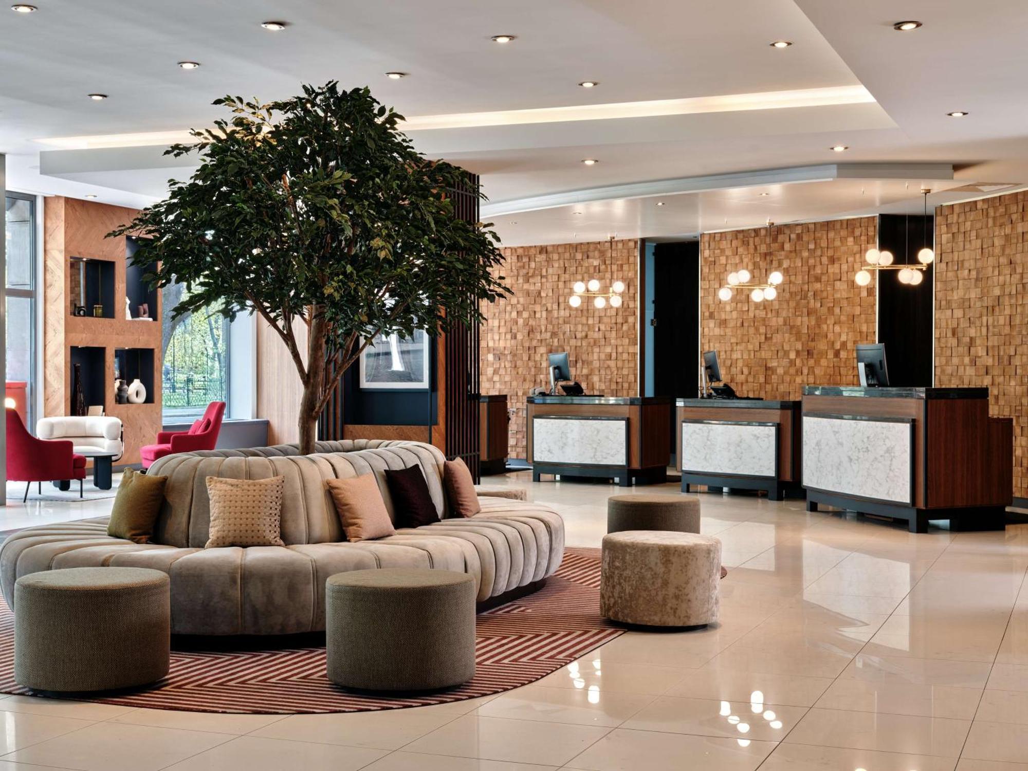 Radisson Blu Hotel Krakow Exterior photo The photo shows a modern hotel lobby. The space features a circular, plush seating area in neutral tones, surrounded by small, round stools. A large decorative tree adds a touch of greenery to the room. In the background, there are multiple reception