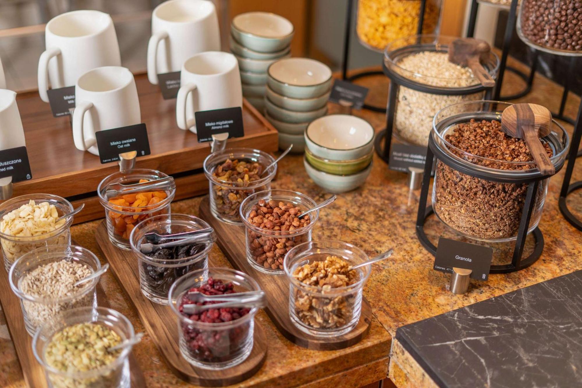 Radisson Blu Hotel Krakow Exterior photo The photo displays a breakfast or snack buffet setup with an assortment of items. There are several white mugs and smaller bowls on a wooden tray, indicating that beverages may be served. In the foreground, there are various glass containers filled w
